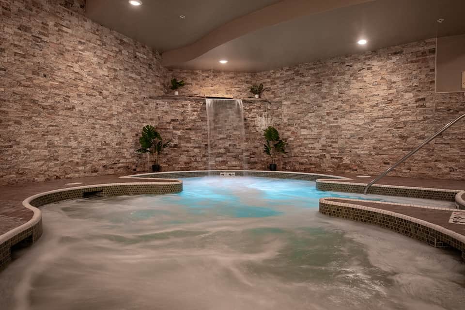 Indoor spa pool with brick walls, waterfall feature, and blue-lit water surrounded by plants