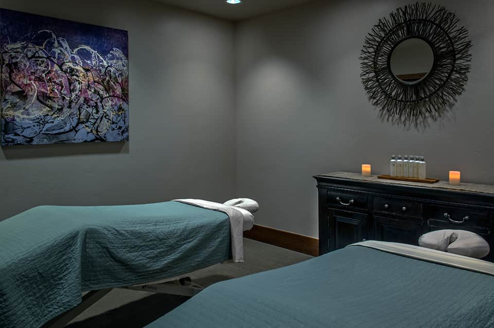 Spa treatment room with two massage beds, teal bedding, artwork, and peaceful ambiance