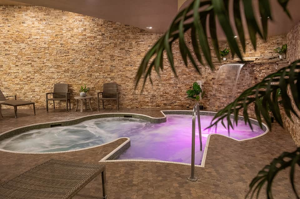 Indoor spa with illuminated purple jacuzzi, soaking tub, brick walls, and lounge seating