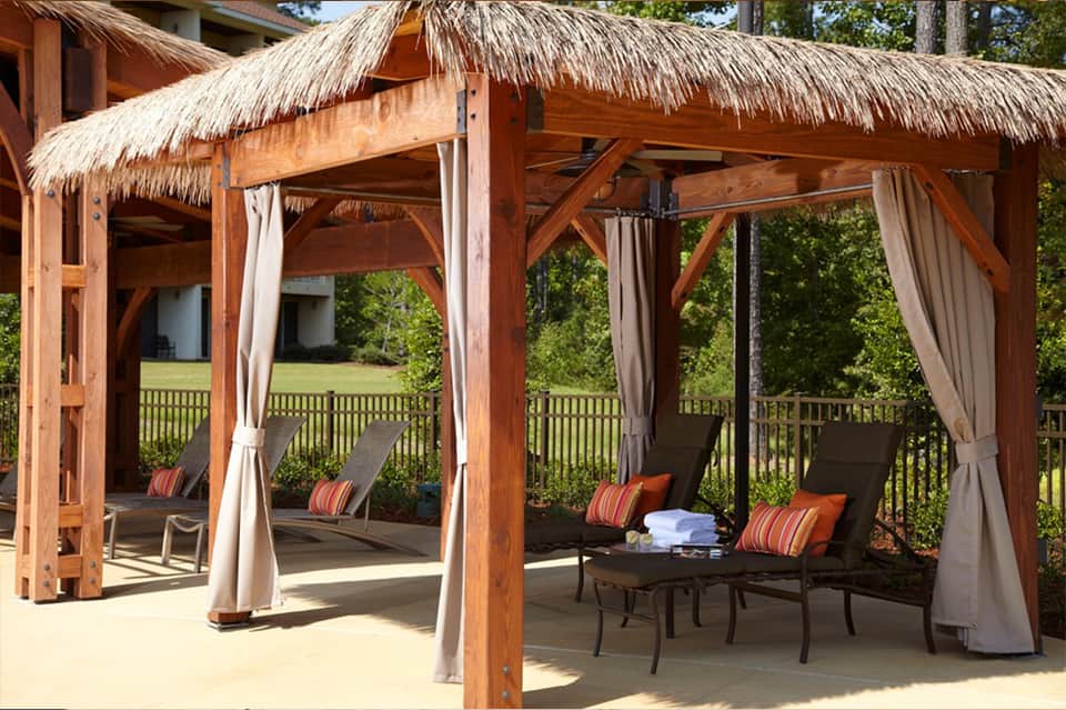 Wooden gazebo with thatched roof, white curtains, and lounge seating overlooking golf course and greenery