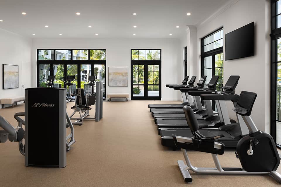 Bright fitness center with cardio equipment, treadmills, exercise bikes, and large windows overlooking outdoor areas