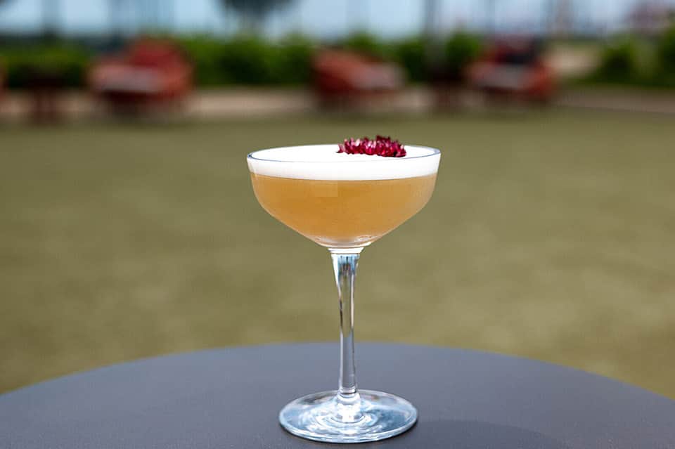 Craft cocktail with foam and garnish served outdoors with blurred water background