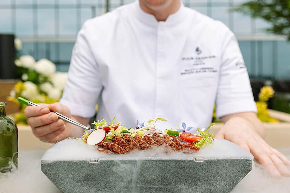Chef in white jacket presenting plated dish with microgreens and garnish outdoors