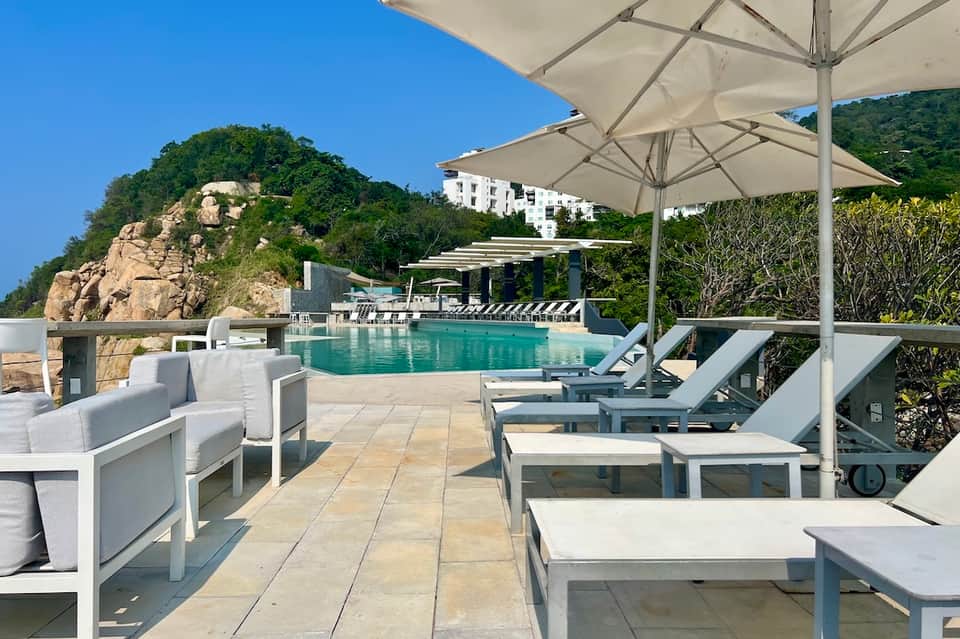 Cliffside pool area with white lounge chairs, large umbrellas, and mountain-ocean landscape views