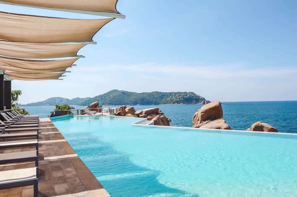 Infinity pool with ocean and mountain views, sun sails, loungers, and natural rock formations