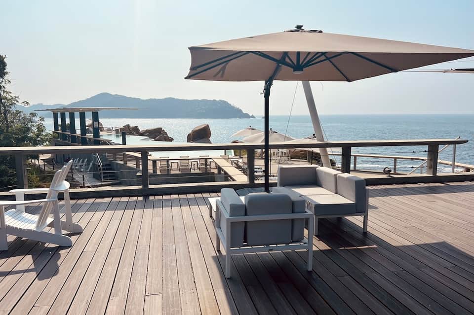 Coastal deck overlooking ocean with white lounge seating, large umbrella, wooden flooring, and island views