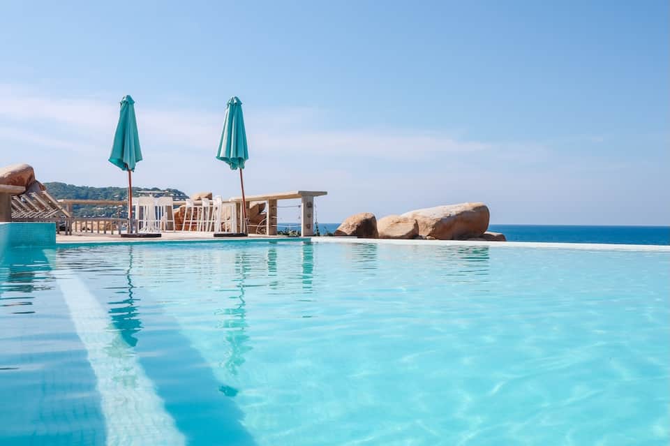 Infinity pool overlooking ocean with teak loungers, turquoise umbrellas, and rocky landscape