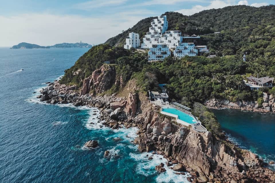 Clifftop resort with white architecture perched on rocky coastline overlooking turquoise ocean and lush hillside vegetation