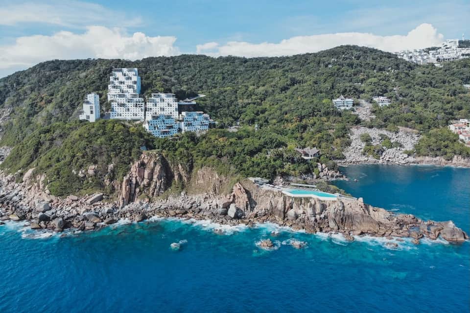 Aerial view of clifftop resort buildings nestled in forested hillside overlooking turquoise ocean waters
