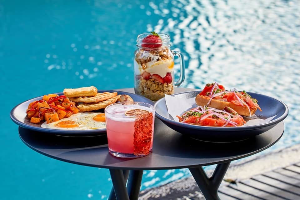 Colorful poolside dining setup with breakfast plate, sandwich, beverage, and granola parfait on dark table