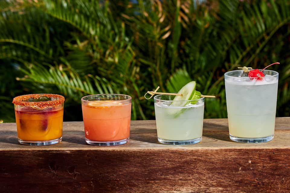 Four colorful tropical cocktails on wooden bar counter with greenery background