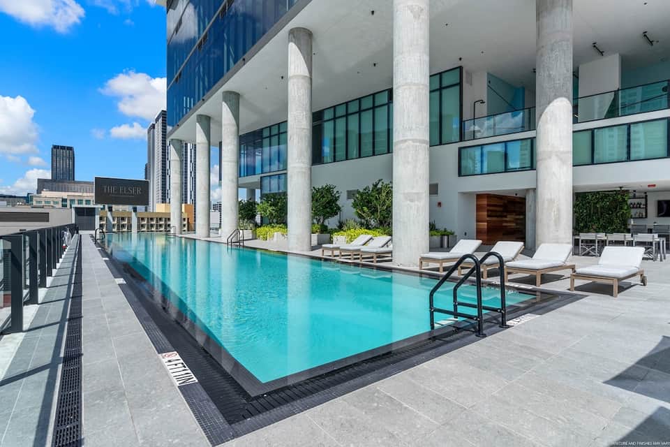 Modern rooftop pool with turquoise water, white lounge chairs, concrete columns, and city skyline views