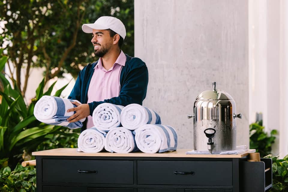 Staff member with rolled towels and beverage dispenser in outdoor garden spa setting