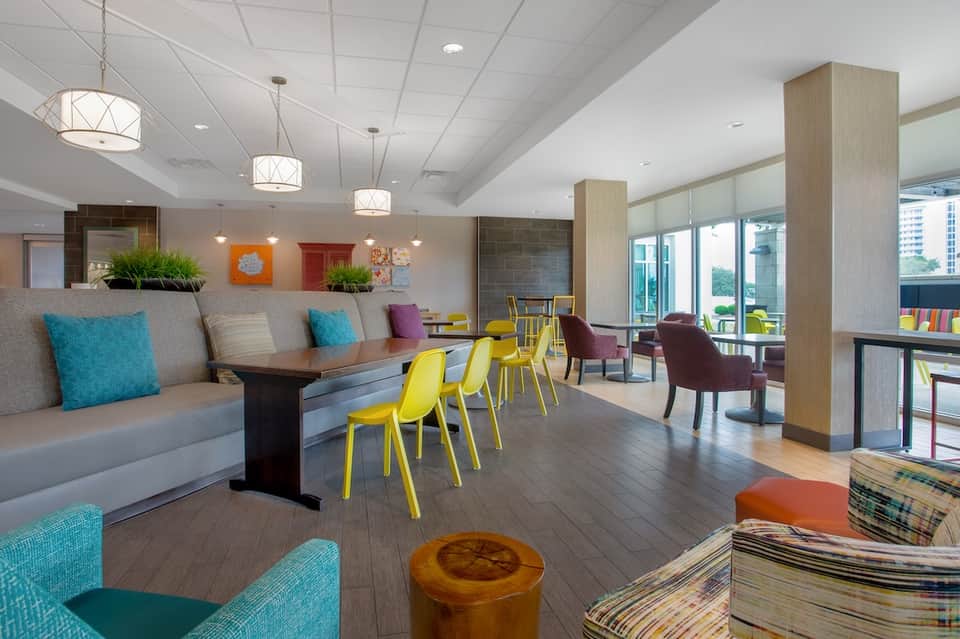 Contemporary hotel lobby with yellow chairs, gray seating, colorful accents, pendant lights, and large windows