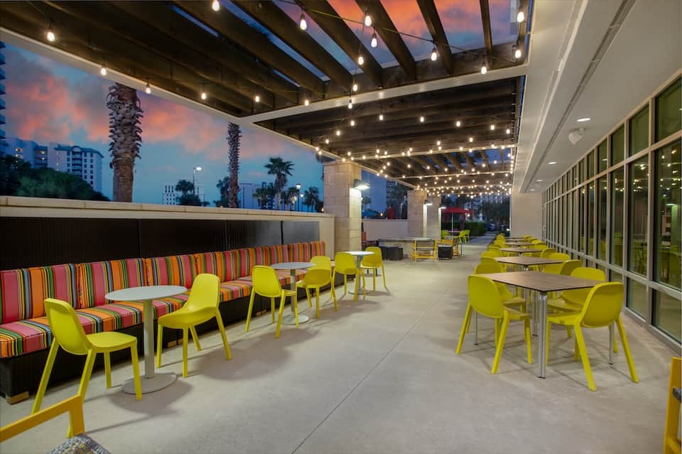 Rooftop terrace at dusk with striped seating, yellow chairs, string lights, palm trees, and sunset sky