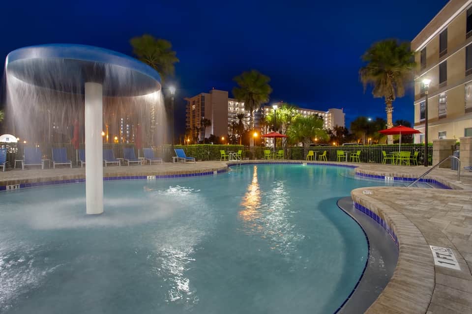 Illuminated resort pool at night with water feature, palm trees, and hotel buildings
