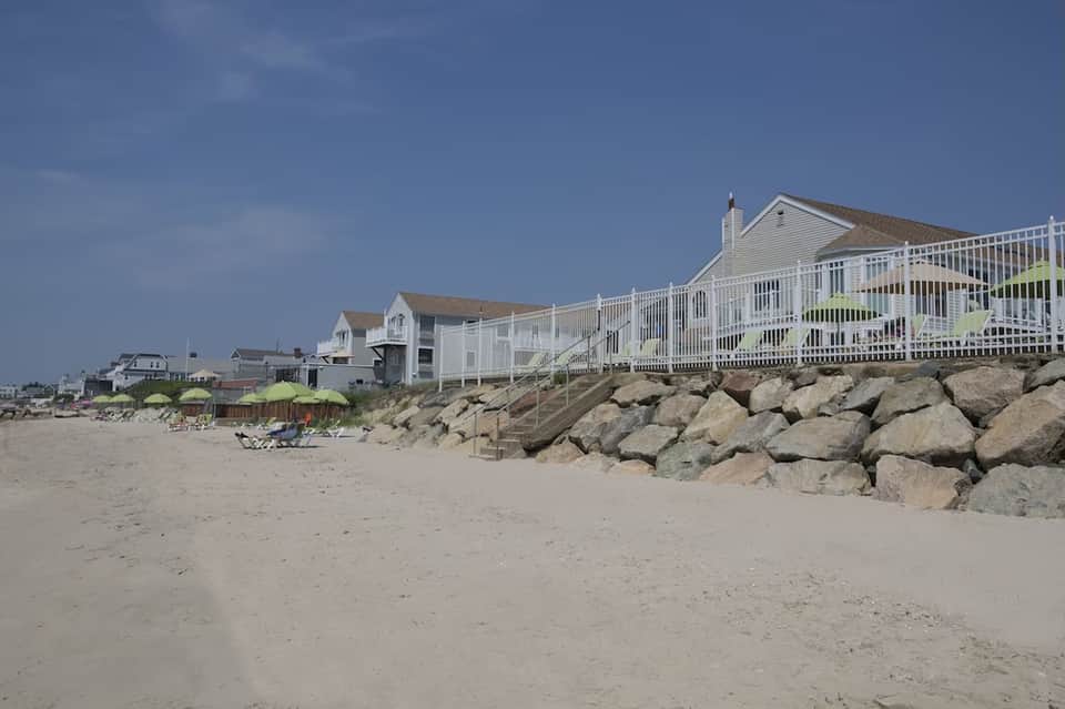 Beachfront homes with white railings on sandy shore protected by large rock breakwater