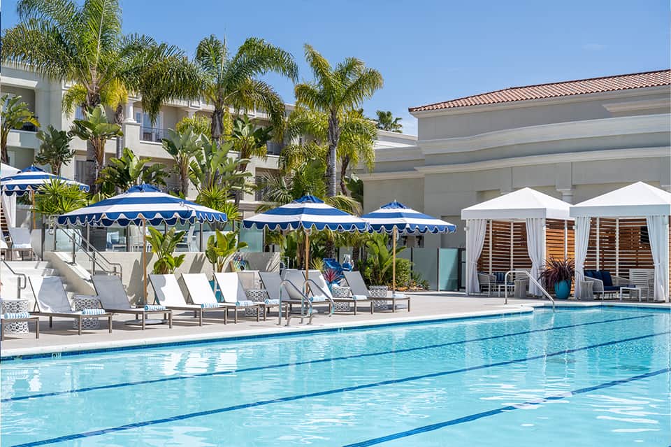 Outdoor lap pool with blue and white striped umbrellas, white loungers, palm trees, and modern buildings
