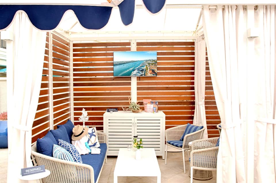 Private poolside cabana with wooden slats, navy awning, white curtains, and woven chairs