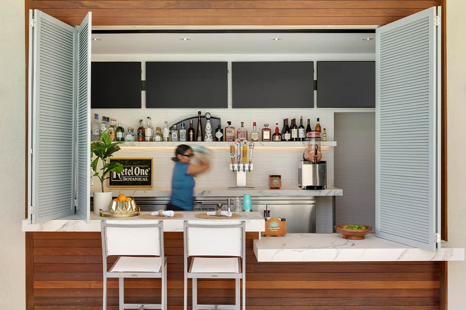 Modern home bar with white shutters, spirit bottles on shelving, and two white chairs at marble counter