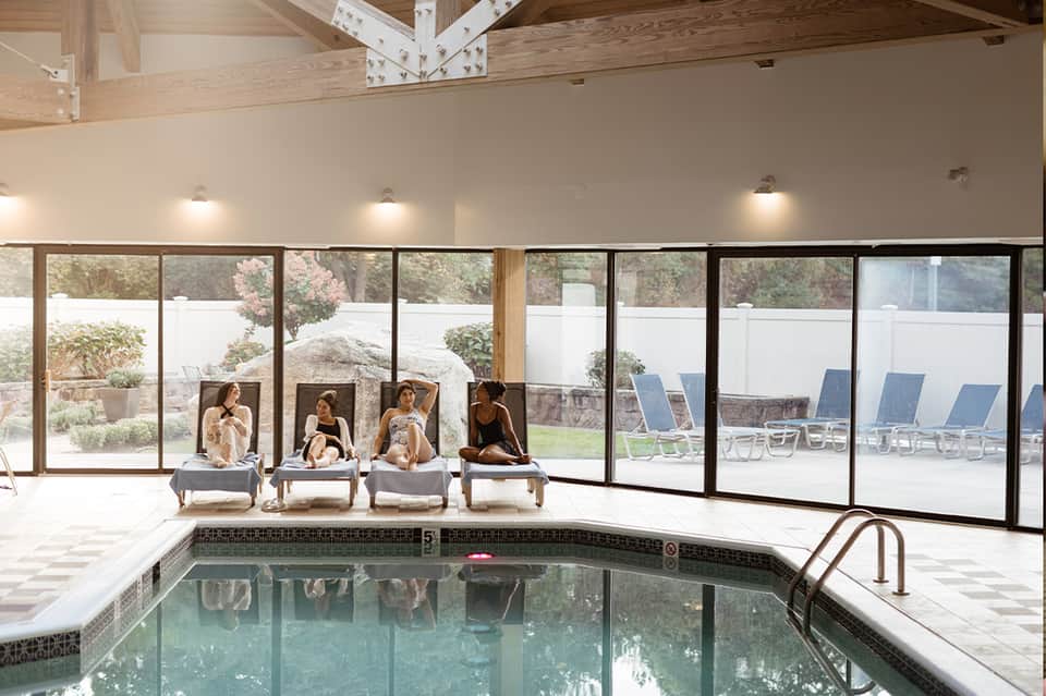 Indoor pool area with lounge chairs, large windows overlooking landscape, and exposed wooden beams
