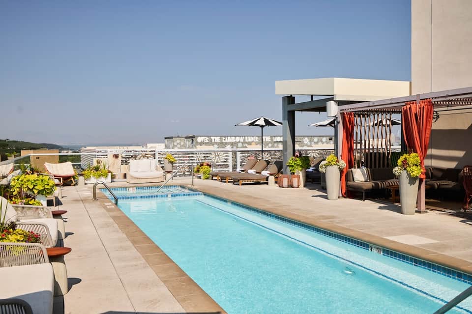 Rooftop pool deck with lap pool, lounge chairs, umbrellas, orange curtains, and city skyline views