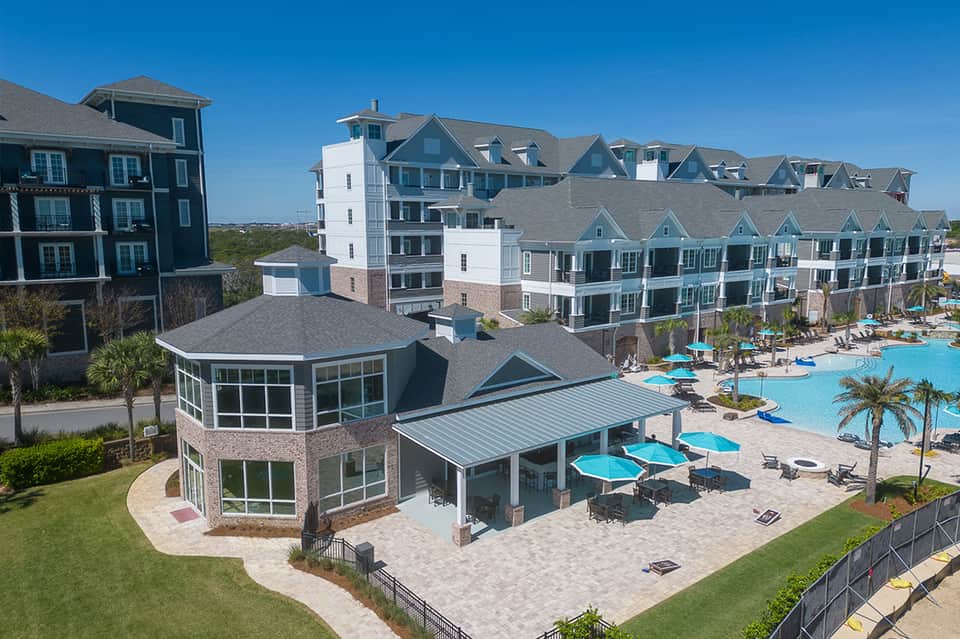 Aerial view of beachfront resort with multi-story buildings, pool, blue umbrellas, and sandy beach