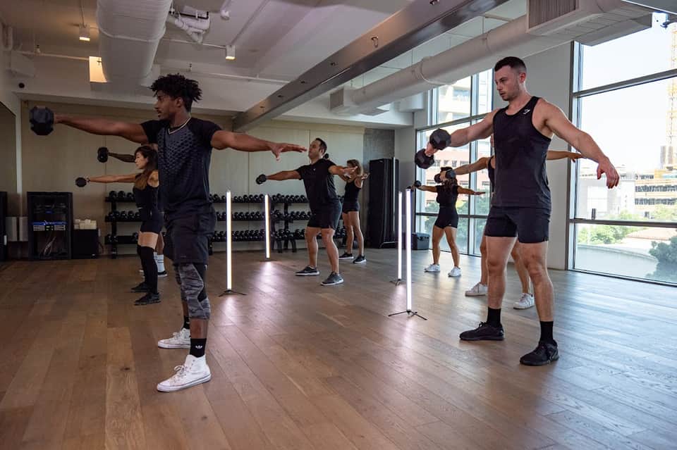 Fitness class in progress with multiple people holding dumbbells in bright gym with large windows