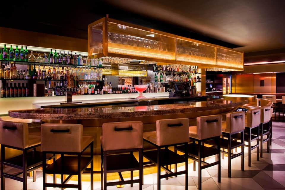 Upscale bar interior with illuminated shelving, copper stools, and ambient lighting