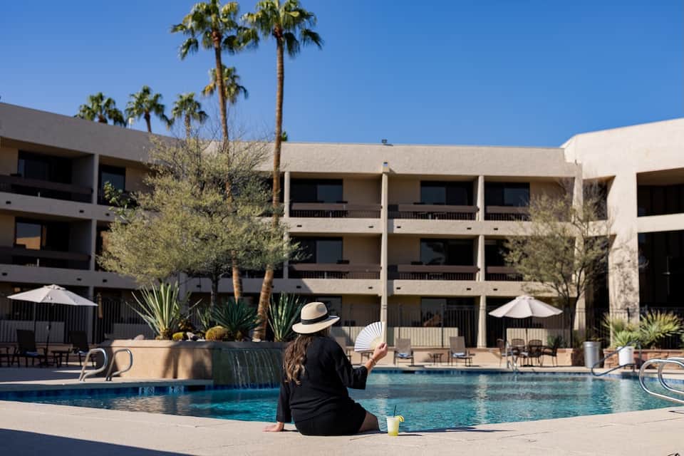 Woman in black shirt and hat sitting on edge of resort pool facing modern concrete building with balconies