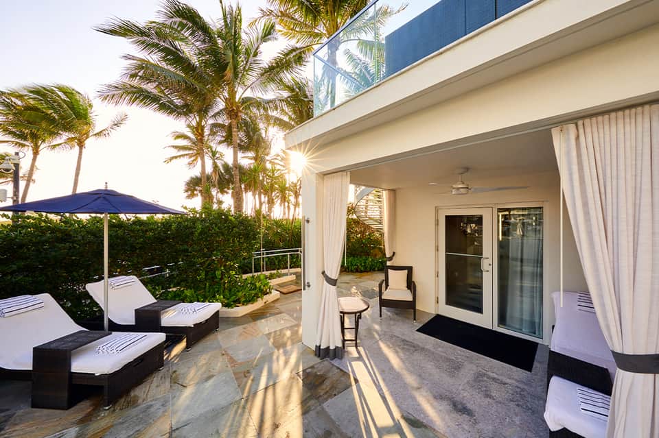 Luxury resort patio with lounge chairs, blue umbrella, palm trees, ocean view, and coastal architecture
