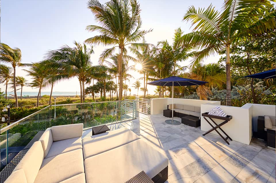 Oceanfront terrace with white lounge seating, blue umbrellas, palm trees, and coastal landscape view