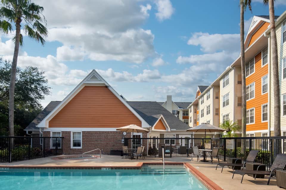 Resort pool with brick clubhouse, lounge chairs, palm trees, and colorful residential buildings