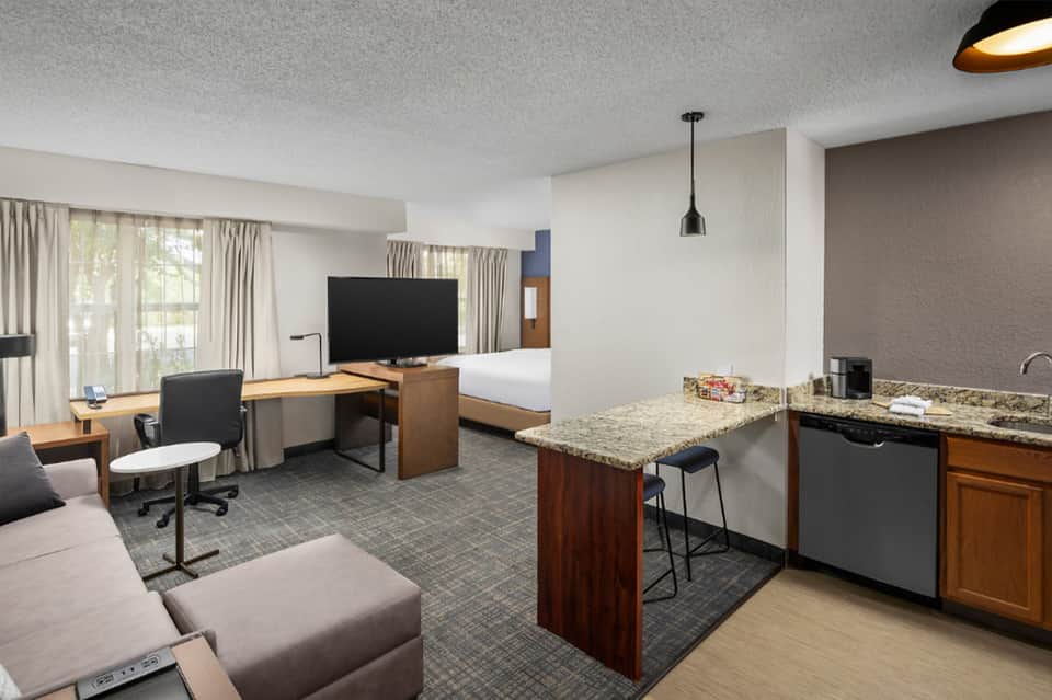 Hotel suite with kitchenette, work desk, TV, and bed visible in background with windows