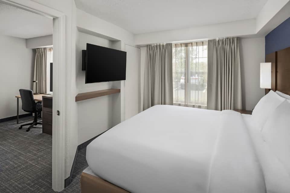 Hotel guest room with white bedding, wall-mounted TV, work desk, chair, and large windows with neutral curtains