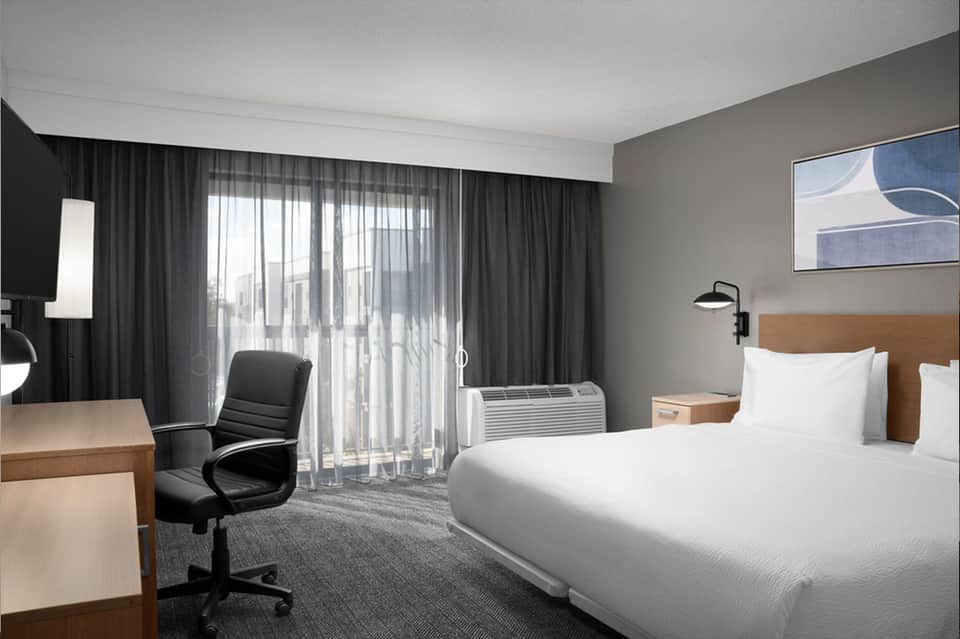 Modern hotel bedroom with queen bed, work desk, balcony access, and contemporary furnishings