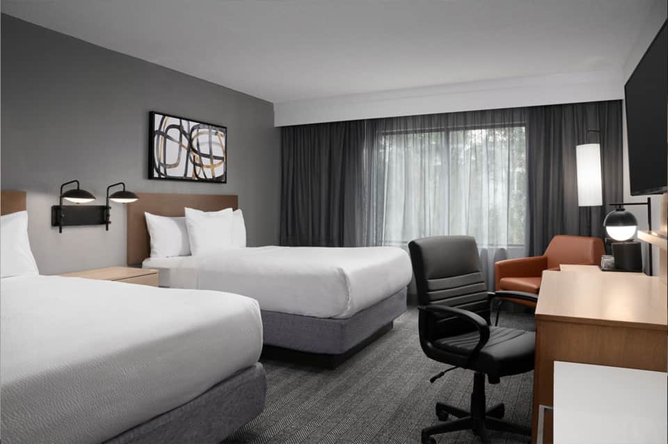 Modern hotel room with two queen beds, work desk, sleek furnishings, and floor-to-ceiling windows.