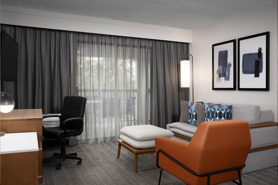 Modern hotel suite living area with orange accent chair, sofa, desk, and floor-to-ceiling windows