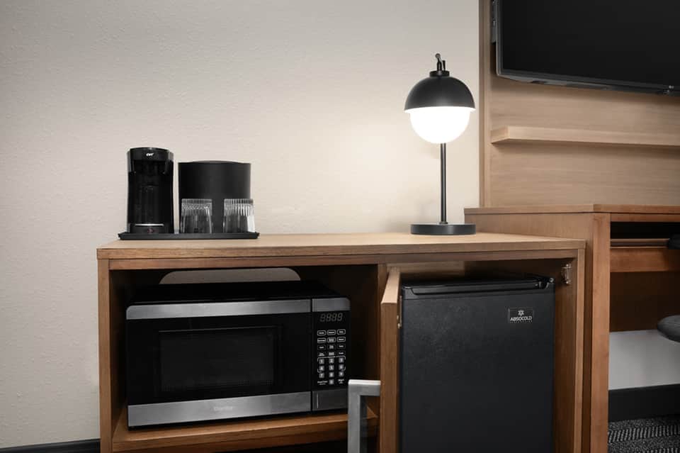 Hotel room desk with coffee maker, mini-fridge, desk lamp, and TV mounted on wooden wall unit
