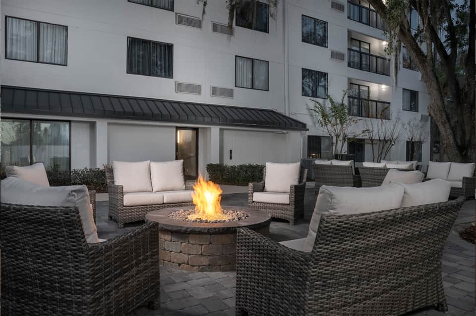 Outdoor fire pit lounge area with wicker seating and cushions in front of modern hotel building