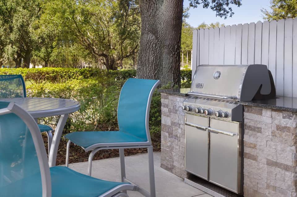 Outdoor patio with turquoise dining chairs, gas grill, white fence, large tree, and manicured landscaping.
