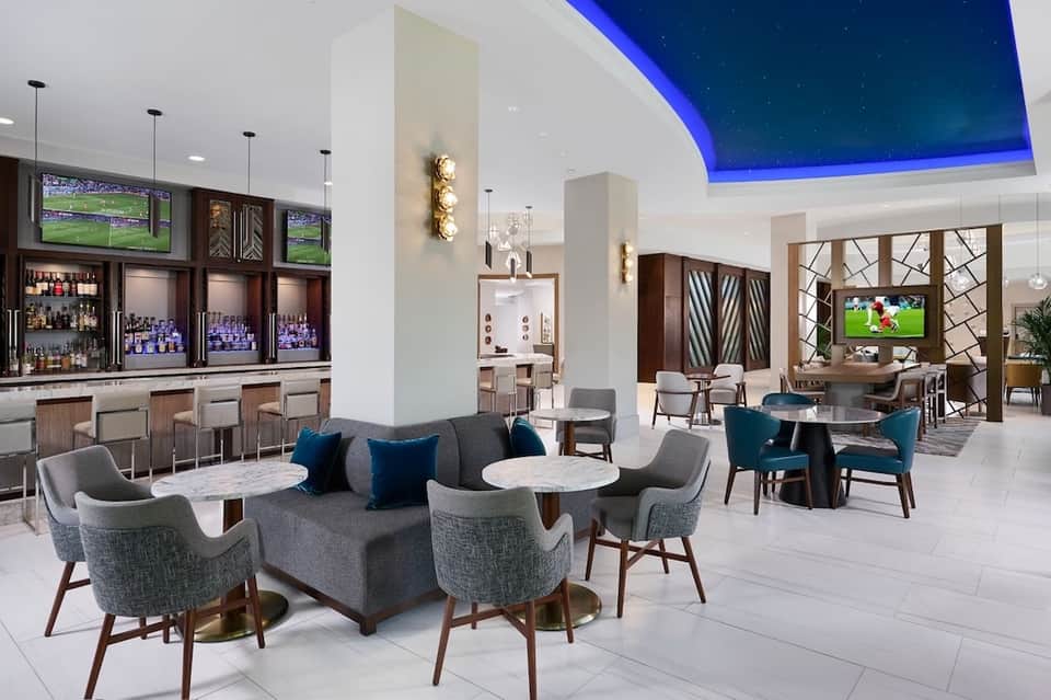 Modern hotel lobby lounge with blue-lit ceiling, contemporary furniture, bar, and flat-screen TVs