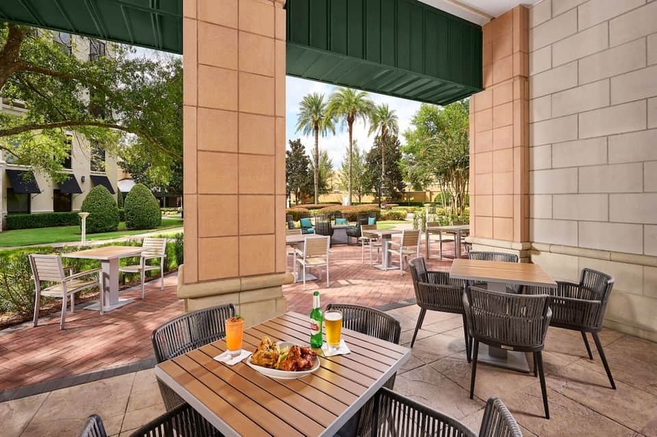 Covered outdoor patio with wooden tables, rope chairs, beverages, and view of tropical landscaping