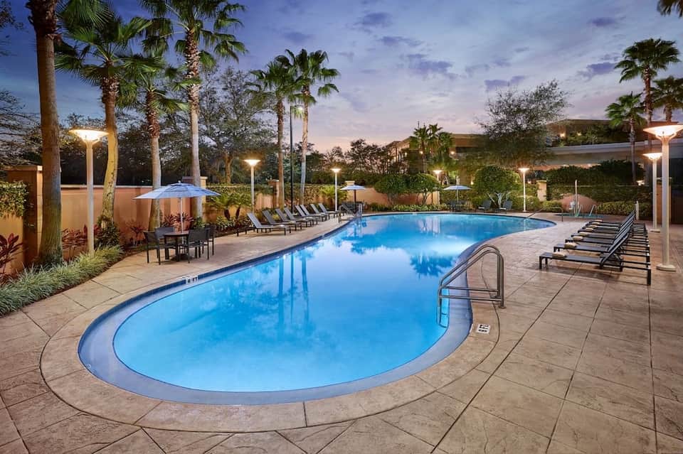 Illuminated resort pool at dusk with palm trees, umbrellas, lounge chairs, and landscaping