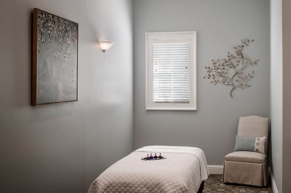 Minimalist spa treatment room with massage table, neutral walls, window with shutters, and botanical wall art