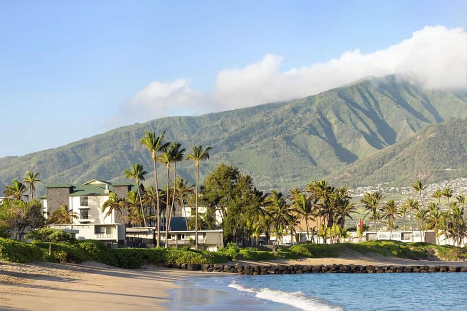 Beachfront resort with white buildings, palm trees, and mountains rising behind sandy beach