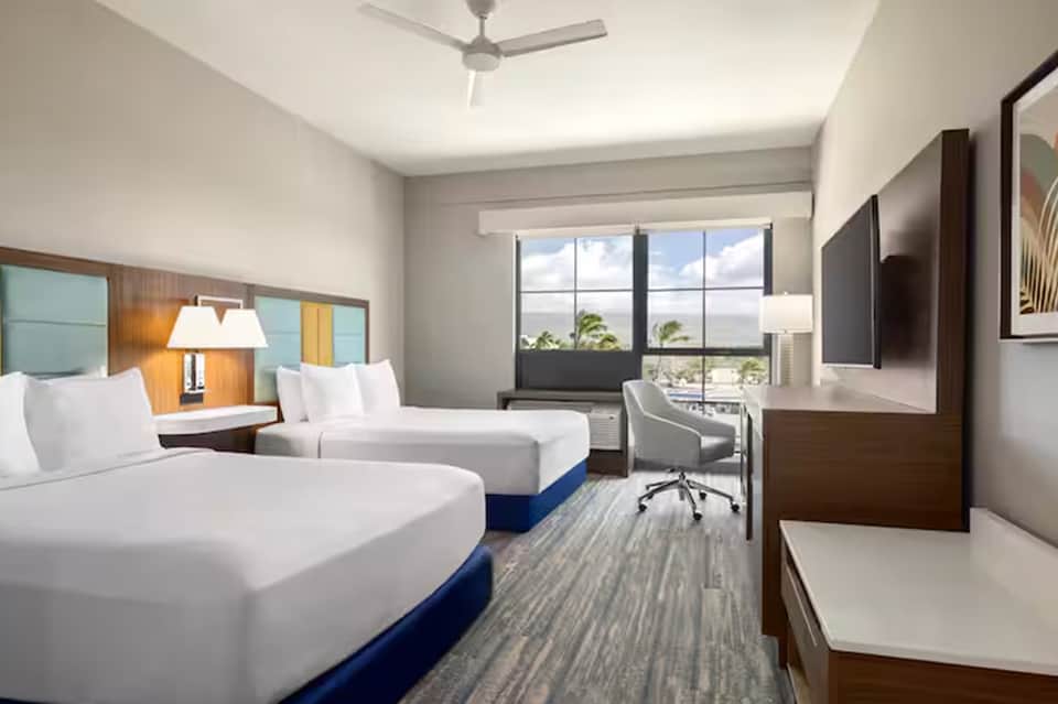 Hotel room with two queen beds, ocean view, desk area, and TV mounted on wall