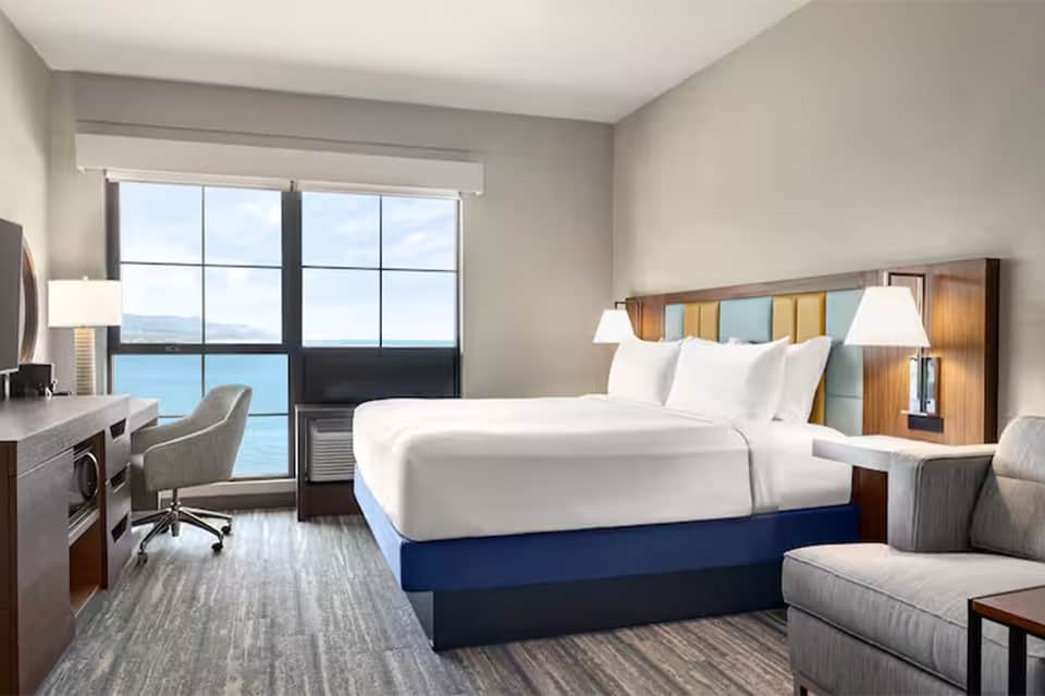 Modern hotel room with ocean view, king bed with blue base, desk, and floor-to-ceiling windows