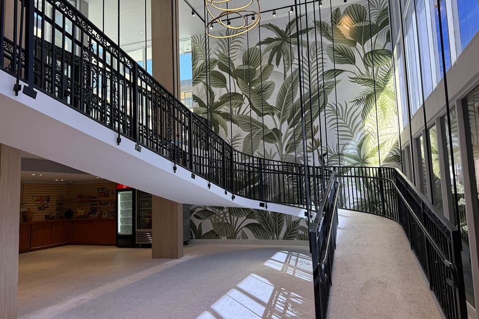 Modern hotel lobby with two-story atrium, botanical wall mural, and curved balcony with black railing