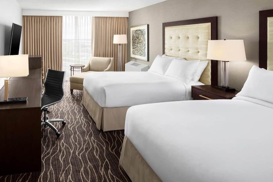 Hotel bedroom with two queen beds, beige headboards, dark wood furniture, and large windows